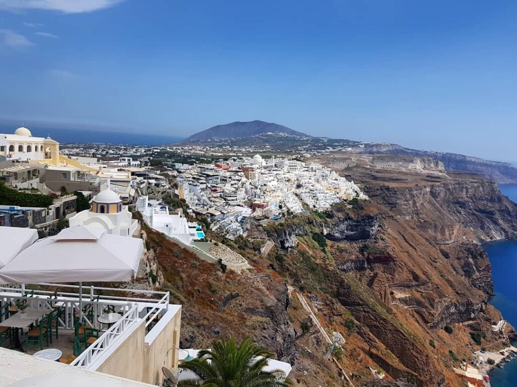 Holland America Line, Cruise Travelers, Koningsdam, Thira, Santorini, Greece | Luxury catamaran cruising through Santorini’s volcanic caldera, showcasing whitewashed buildings perched on cliffs, a secluded cove with warm mineral-rich waters, and guests enjoying a stunning sunset while savoring local delicacies prepared by the onboard chef during an exclusive sailing experience.