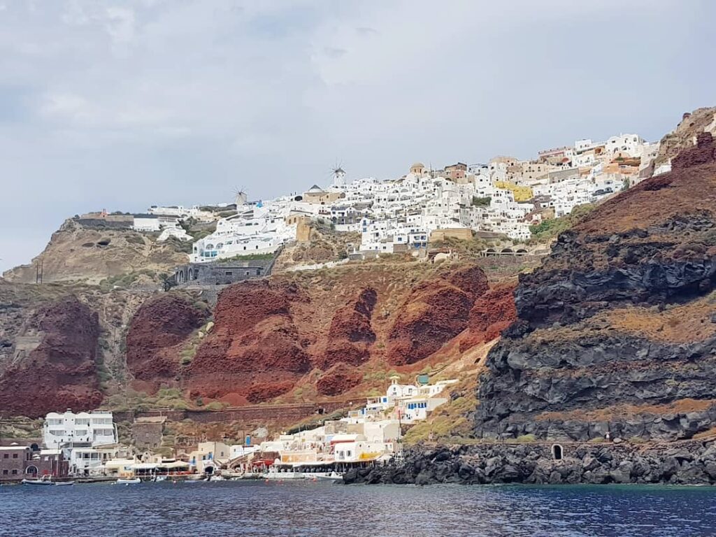 Holland America Line, Cruise Travelers, Koningsdam, Thira, Santorini, Greece | Luxury catamaran cruising through Santorini’s volcanic caldera, showcasing whitewashed buildings perched on cliffs, a secluded cove with warm mineral-rich waters, and guests enjoying a stunning sunset while savoring local delicacies prepared by the onboard chef during an exclusive sailing experience.