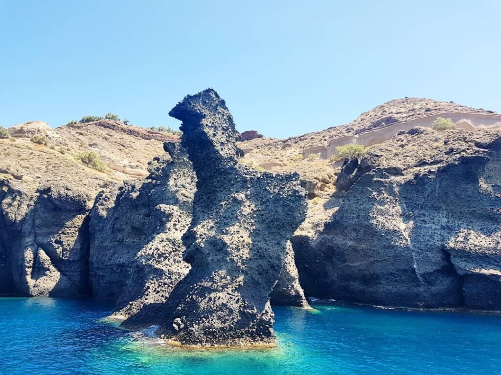 Holland America Line, Cruise Travelers, Koningsdam, Thira, Santorini, Greece | Luxury catamaran cruising through Santorini’s volcanic caldera, showcasing whitewashed buildings perched on cliffs, a secluded cove with warm mineral-rich waters, and guests enjoying a stunning sunset while savoring local delicacies prepared by the onboard chef during an exclusive sailing experience.