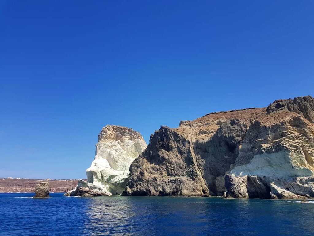 Holland America Line, Cruise Travelers, Koningsdam, Thira, Santorini, Greece | Luxury catamaran cruising through Santorini’s volcanic caldera, showcasing whitewashed buildings perched on cliffs, a secluded cove with warm mineral-rich waters, and guests enjoying a stunning sunset while savoring local delicacies prepared by the onboard chef during an exclusive sailing experience.