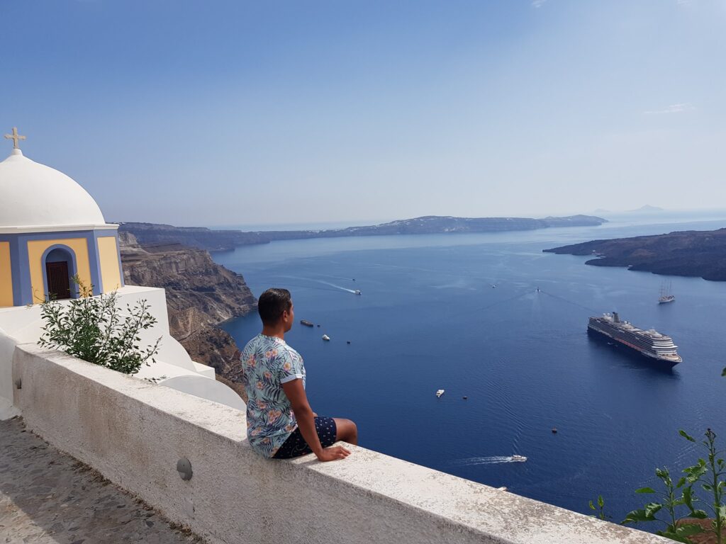 Holland America Line, Cruise Travelers, Koningsdam, Thira, Santorini, Greece | Luxury catamaran cruising through Santorini’s volcanic caldera, showcasing whitewashed buildings perched on cliffs, a secluded cove with warm mineral-rich waters, and guests enjoying a stunning sunset while savoring local delicacies prepared by the onboard chef during an exclusive sailing experience.